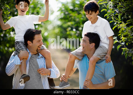 Zwei lächelnde Mitte - erwachsene Männer, die ihre jungen Söhne tragen auf ihren Schultern die über einen Apple Orchard in der Sonne. Stockfoto