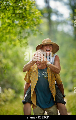 Lächelnd reifer Mann, der seine Frau ein huckepack Fahrt, während sie seine Augen mit den Händen umfasst. Stockfoto