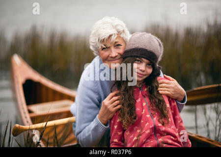 Reife Frau umarmt ihre Enkelin, als sie zusammen sitzen und sich für ein Portrait in einem hölzernen Kanu zwischen Schilf an einem Seeufer darstellen. Stockfoto
