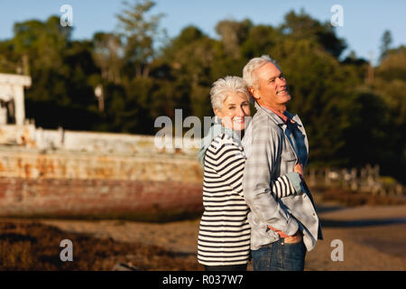 Älteres Ehepaar mit ihren Armen um einander stehen auf einem Strand vor einem verlassenen Boot. Stockfoto