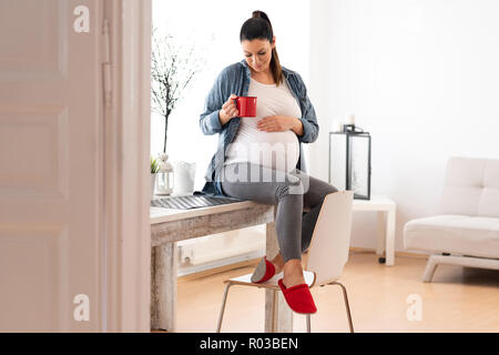 Ein schönes Lächeln schwangere Frau sitzt auf einem Küchentisch in einer Jeansjacke und trinken einen Kaffee aus einer roten Tasse. Stockfoto