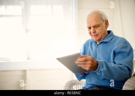 Gerne älterer Mann Spaß suchen an einem digitalen tablet, während er sich in einem Sessel sitzt. Stockfoto