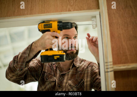 Mitte der erwachsene Mann konzentrieren, als er eine elektrische Bohrmaschine hält, während das Ausbessern der Türrahmen. Stockfoto