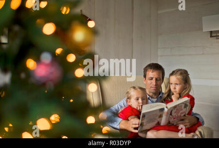 Vater ein Buch lesen zu seinen zwei Töchtern neben dem Weihnachtsbaum. Stockfoto