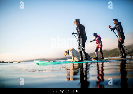 Zwei Männer und ein junges Mädchen paddleboarding auf einem See mit ihrem Hund. Stockfoto