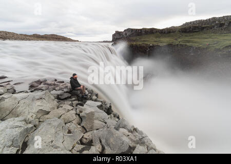 Mann sitzt auf der Klippe neben dem Wasserfall Dettifoss in Vatnayokull National Park, Island. Bewölkten Tag, langen Belichtungszeit. Stockfoto