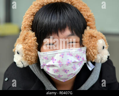 Junge Taiwanese Frau mit lächelnden Augen trägt ein Einmal-OP-ohrläppchen Gesichtsmaske und flauschige winter Ohrenschützer. Stockfoto