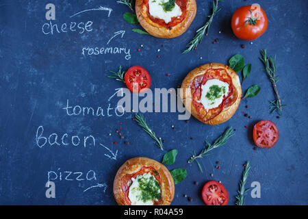 Drei hausgemachten Mini Pizza mit Tomaten, Käse und Speck, Verletzungen und Gewürze auf einem dunklen Hintergrund mit einer schriftlichen Rezept. Stockfoto