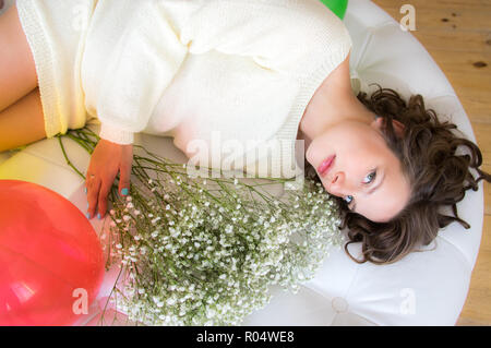 Schwangere Frau in weißem Kleid liegt auf runden Sofa mit Blumenstrauß Stockfoto