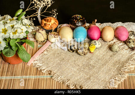 Bemalte Ostereier und Wachteln Eier, Gebäck, eine Kanne Weiß Violett, Spielzeug gelben Küken mit sackleinen auf einem Stroh Teppich Stockfoto