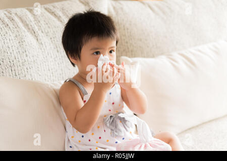 Kranken kleinen asiatischen Mädchen wischen oder Reinigung der Nase mit einem Taschentuch, auf dem Sofa zu Hause sitzen. Medizin und Gesundheit Konzept. Stockfoto