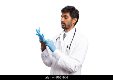 Indische Arzt männlichen Blick auf Hand mit konzentrierten Ausdruck als das Setzen auf Blau chirurgische latex Handschuh isoliert auf weißem Stockfoto