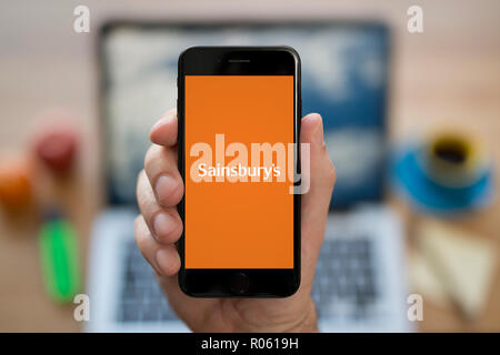 Ein Mann schaut auf seinem iPhone die zeigt das Logo des Sainsbury, während saß an seinem Computer Schreibtisch (nur redaktionelle Nutzung). Stockfoto