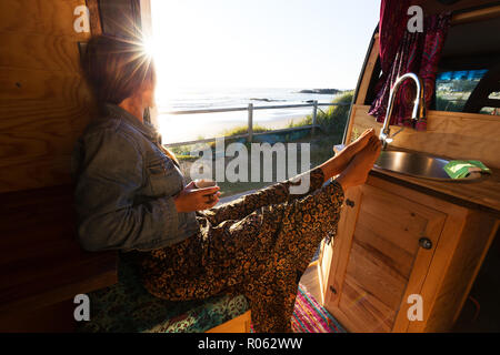 Junge Frau im Wohnmobil, van Life Theme bei Sonnenaufgang am Strand Stockfoto