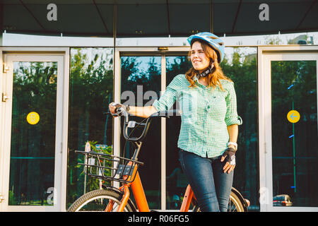 Thema auf dem Fahrrad zur Arbeit. Eine junge Kaukasier Frau auf umweltfreundliche Verkehrsmittel Fahrrad im Büro angekommen. Mädchen in ein Fahrrad parken Büro Stockfoto