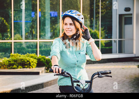 Thema auf dem Fahrrad zur Arbeit. Eine junge Kaukasier Frau auf umweltfreundliche Verkehrsmittel Fahrrad im Büro angekommen. Mädchen in ein Fahrrad parken Büro Stockfoto
