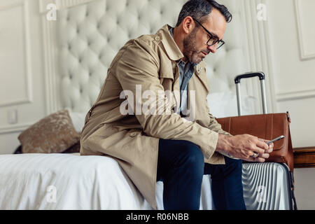 Portrait von Reifen Geschäftsmann im Hotelzimmer zu sitzen. Mann in formale Abnutzung sitzen auf dem Bett mit Handy. Stockfoto