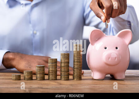 Afrikanischer Mann einfügen Münze in Sparschwein in der Nähe von Stapel von Münzen Stockfoto