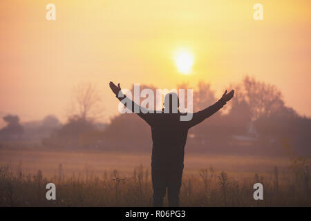 Ein glücklicher Mann mit den Händen in der Luft stehend auf dem Feld in den frühen Morgenstunden und mit Blick auf einen Sonnenaufgang Stockfoto