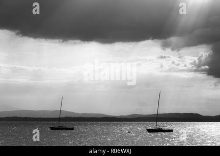 Ein paar leer, kleinen Segelboot auf einem See, unter einem Moody Himmel mit Sonnenstrahlen durch Filterung. Stockfoto