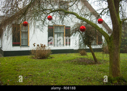 Amersfoort, Niederlande - 2018-12-16: rote Christbaumkugeln als Dekoration in Weiß lackiert Bauernhaus mit Strohdach und Fensterläden Stockfoto