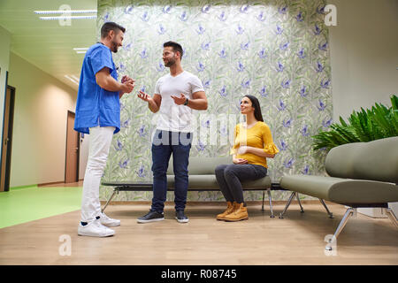 Ein schöner männlicher Arzt Beratung mit einem Jungen schwanger Paar in den ärzten Wartezimmer. Stockfoto