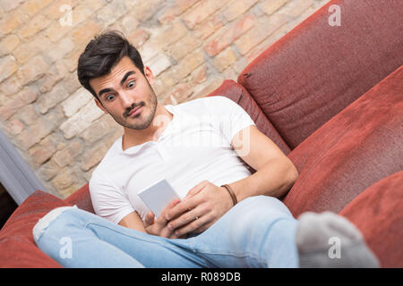 Ein überrascht hübscher junger Mann in einem weißen T-Shirt Festlegung auf ein Sofa und mit seinem Smartphone. Stockfoto