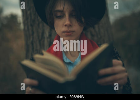 Junge Frau mit Buch in der Hand sitzt neben Baumstamm und liest während der Dämmerung. Stockfoto