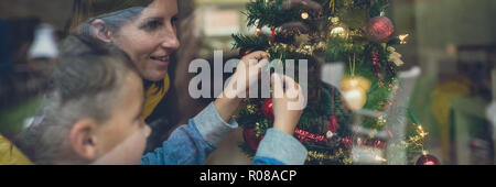 In der breiten Ansicht Bild von Mutter und Sohn schmücken Weihnachtsbaum zusammen. Blick durch ein Fenster mit retro Filter Effekt. Stockfoto
