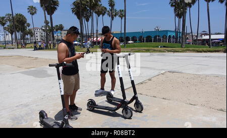 Zwei junge, muskulöse Asiatische Männer nutzen das Handy app zu entsperren und dockless elektrische Vogel Motorroller auf der Venice Beach Radweg mieten Stockfoto