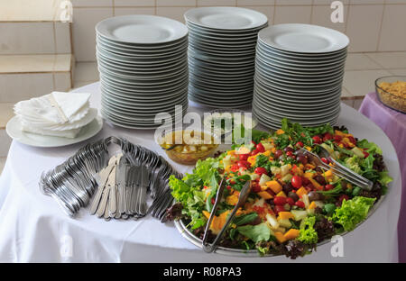 Lecker Salat von Gemüse und Obst. Kopfsalat, Tomaten, Petersilie, Rucola, Traube, Mango, Melone. Auf dem Tisch liegt ein Stapel Teller, Gabeln und Messer. Stockfoto