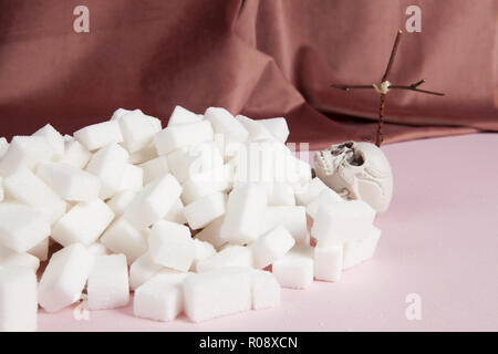 Ein Skelett mit weißen Würfel Zucker wie ein Grab mit einem Kreuz auf einem rosa Hintergrund und vor einem rosa Samtvorhang. Farbharmonie. Minimale Stockfoto