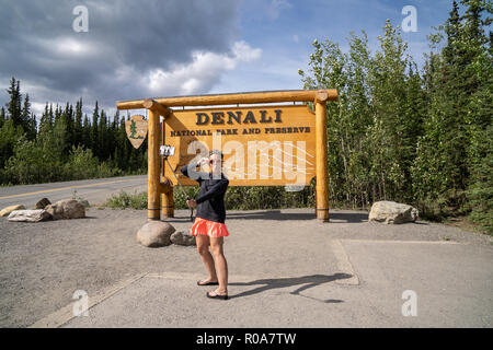 Am 1. August 2018 - DENALI NATIONAL PARK, ALASKA: Ein tourist Frau verwendet eine selfie Stick ein Foto mit Ihrem Smartphone der Park Eingang Schild zu nehmen, Stockfoto