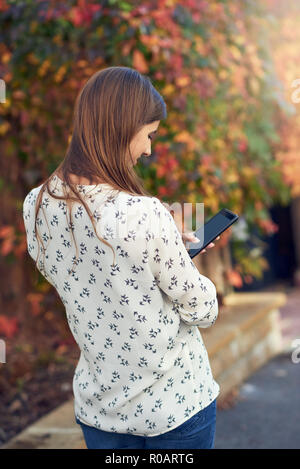 Junge Frau, die in einem Herbst Straße Textnachrichten auf Ihr Mobiltelefon eingeben Eine Nachricht mit dem Rücken zur Kamera und buntes Laub auf einem Kriechgang Stockfoto