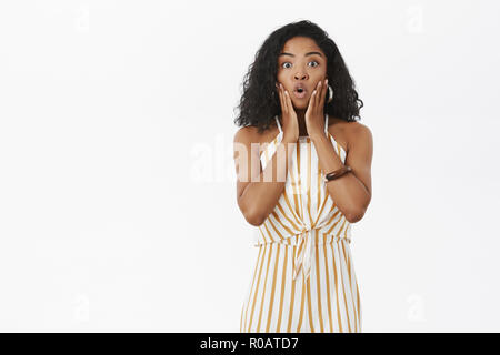 Portrait von überrascht und erstaunt, african american Female in trendigen Sommer Overalls in gelb gestreiften keuchend öffnung öffnung von Erstaunen, Hände auf Gesicht, neugierig und interessiert Stockfoto