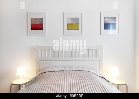Innenraum der Schlafzimmer. Einfach, komfortabel und stilvoll. Lampen Beleuchtung auf ein Bett, Tisch, Bett mit Wolldecke, Dekoration an der Wand drei farbenfrohe Bilder Stockfoto