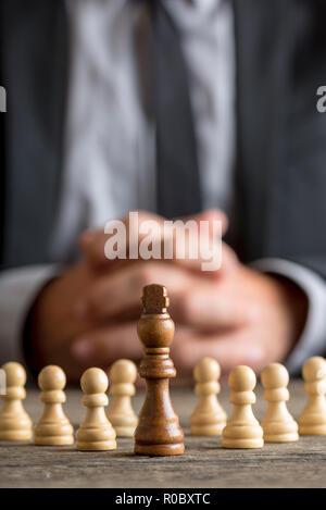 Geschäftsmann mit gefalteten Händen sitzen vor dem dunklen König Schach stehenden Figur unter weißen Bauern auf einem alten Holztisch. Stockfoto