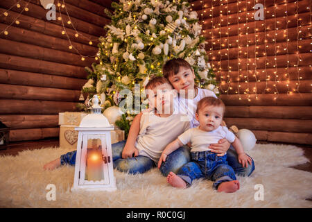 Drei glückliche Kinder - jugendlicher Junge, Kleinkind Mädchen und ihre neugeborenen Bruder - gemeinsam spielen unter einem schönen Weihnachtsbaum. Jungen sitzen Stockfoto