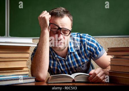 Eine müde und gefoltert zerzaust Student in Gläsern ist schlafen, an einem Tisch mit Stapeln von Büchern vor dem Hintergrund eines kühlen Tafel. Stockfoto