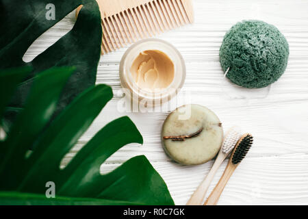Umweltfreundliche natürliche Bambus Zahnbürsten, Zahnpasta, Shampoo bar in Glas, Holz- Bürste und Schwamm konjaku auf weißem Holz mit grünen monstera verlässt. Stockfoto