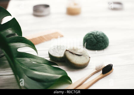 Umweltfreundliche natürliche Bambus Zahnbürsten, Zahnpasta, Shampoo bar in Glas, Holz- Bürste und Schwamm konjaku auf weißem Holz mit grünen monstera verlässt. Stockfoto