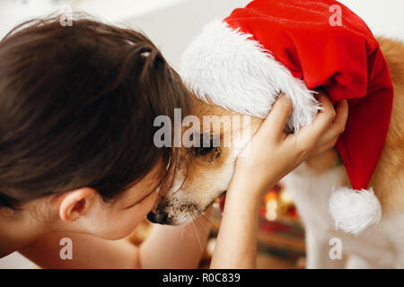 Mädchen umarmt adorable Hund in santa hut mit lustigen Emotionen und Augen, sitzen in festlichen Raum. Frohe Weihnachten Konzept. Süße golden doggy. Atmospheri Stockfoto