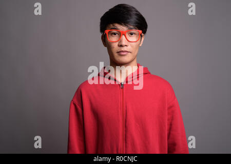 Jungen asiatischen Mann mit roten Mantel mit Brillen gegen Grau Stockfoto
