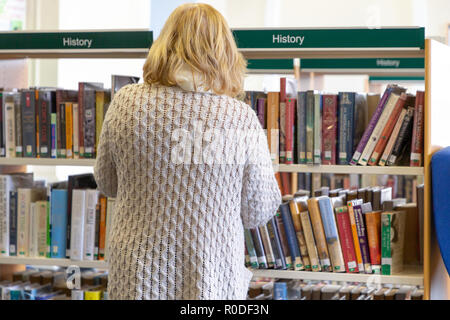 Reife, blonde Dame suchen durch die Bücher in der Geschichte der Bibliothek in Warrington, Cheshire, England, Großbritannien Stockfoto