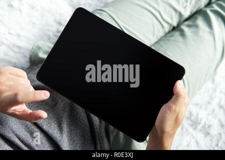 Nahaufnahme von einem jungen Mann bequem auf ein Bett mit einer Tablette und Sie auf den Bildschirm tippen. Halfshot ein Mann sitzt mit schwarzen Bildschirm Tablet in der Hand. Stockfoto