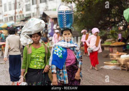 AMARAPURA, Myanmar, 17. JANUAR 2015: Zwei Frauen sind mit einer Tasche und einem Korb auf dem Kopf, zu Fuß in die schmutzige Straße Der Zegyo Markt, in M Stockfoto