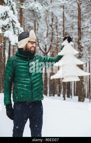 Vertikale Portrait von dlightful positive bärtigen Mann in warmen Winter gekleidet, blickt mit glücklichen Ausdruck an künstlichen Tannenbaum, zeigt Stockfoto