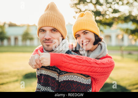 Lächelnd froh, junge weibliche in Hut und warme Pullover aus Baumwolle umfasst ihr Mann, der zurück steht, verbringen Wochenenden zusammen genießen, Spaziergang im Garten o Stockfoto