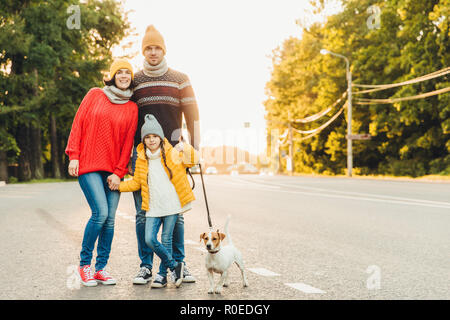 Glückliche Familie tragen Sie warme Kleidung Spaziergang mit Hund auf der Straße, stehen nahe beieinander in der Kamera dar. Kleines Mädchen zeigt ok Zeichen als froh fr zu verbringen Stockfoto