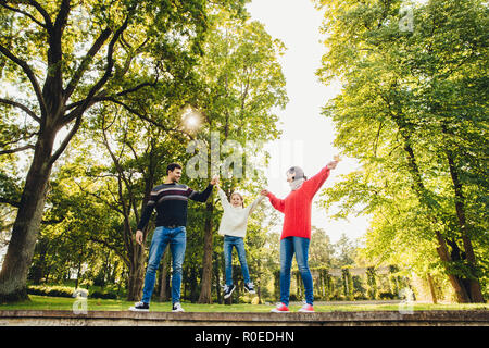 Portrait von freundlichen, liebevollen Familie Spaß haben gemeinsam, freie Zeit draußen verbringen, stand gegen grüne Bäume im Park, versuchen kleine Kind in ha Rock Stockfoto